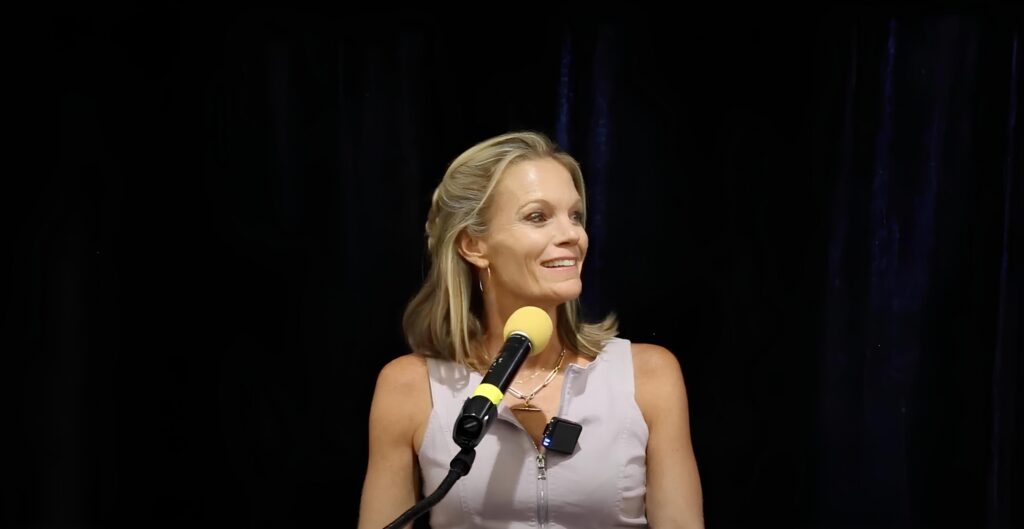 Robyn O'Brien speaking into a yellow covered microphone with a slight smile looking off to the right. She is wearing a purple sleeveless shirt.