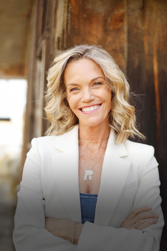Robyn O'Brien standing outdoors in front of a wooden building. She is smiling at the camera and wearing a white blazer jacket and blue camisole with her arms crossed.
